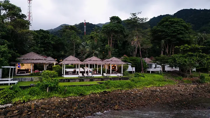 Paradise Cottage vanaf zee: rieten sala's langs de kust met de jungle en de heuvels erachter