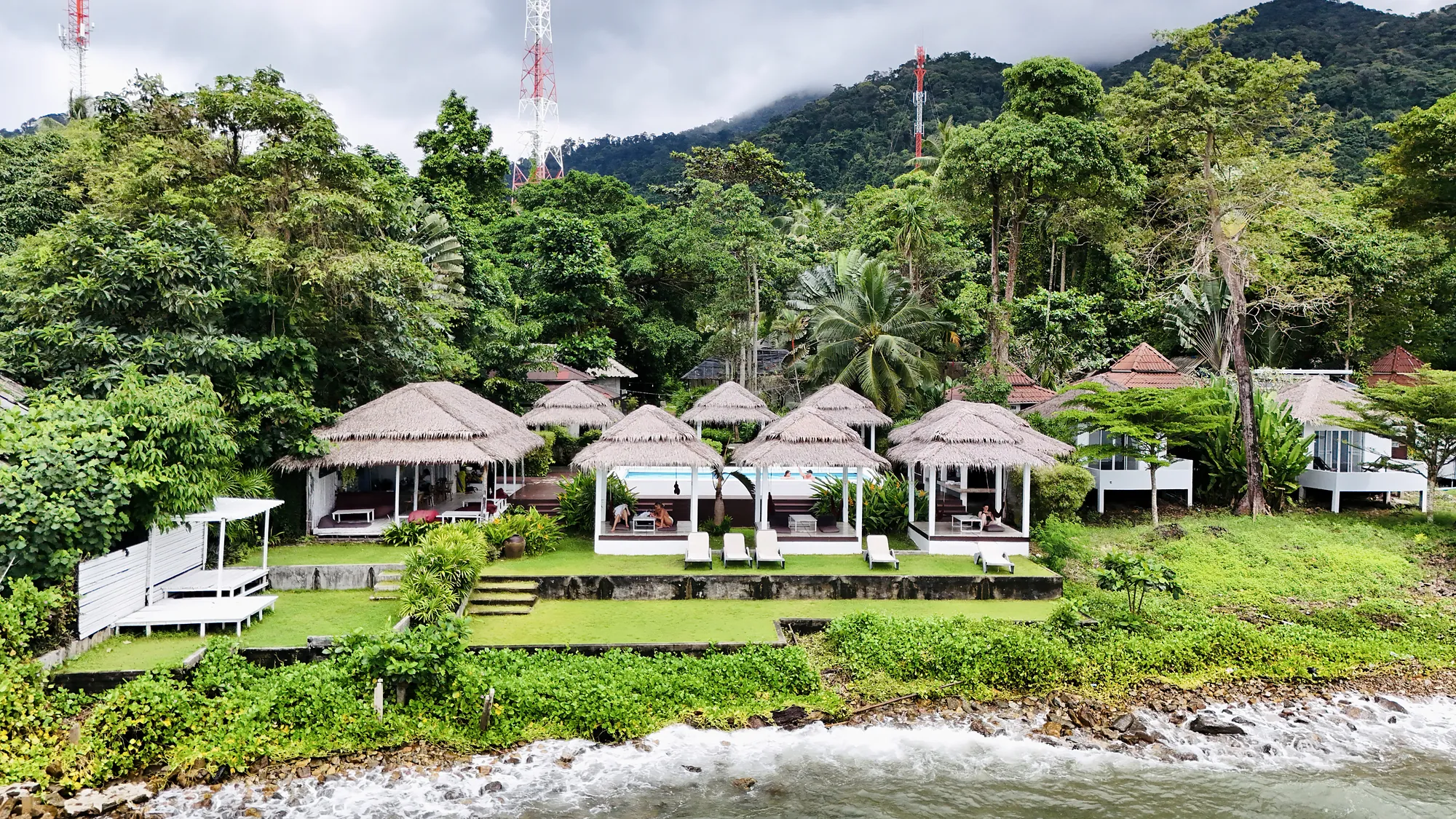 Het resort vanaf het water: golven op de rotsen, het gazon en de rieten sala's onder de jungle
