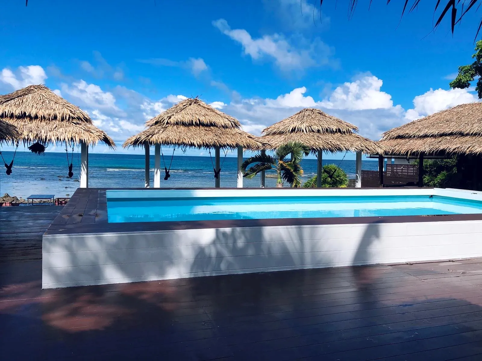 Swimming pool on the wooden deck with thatched salas and the sea under a blue sky